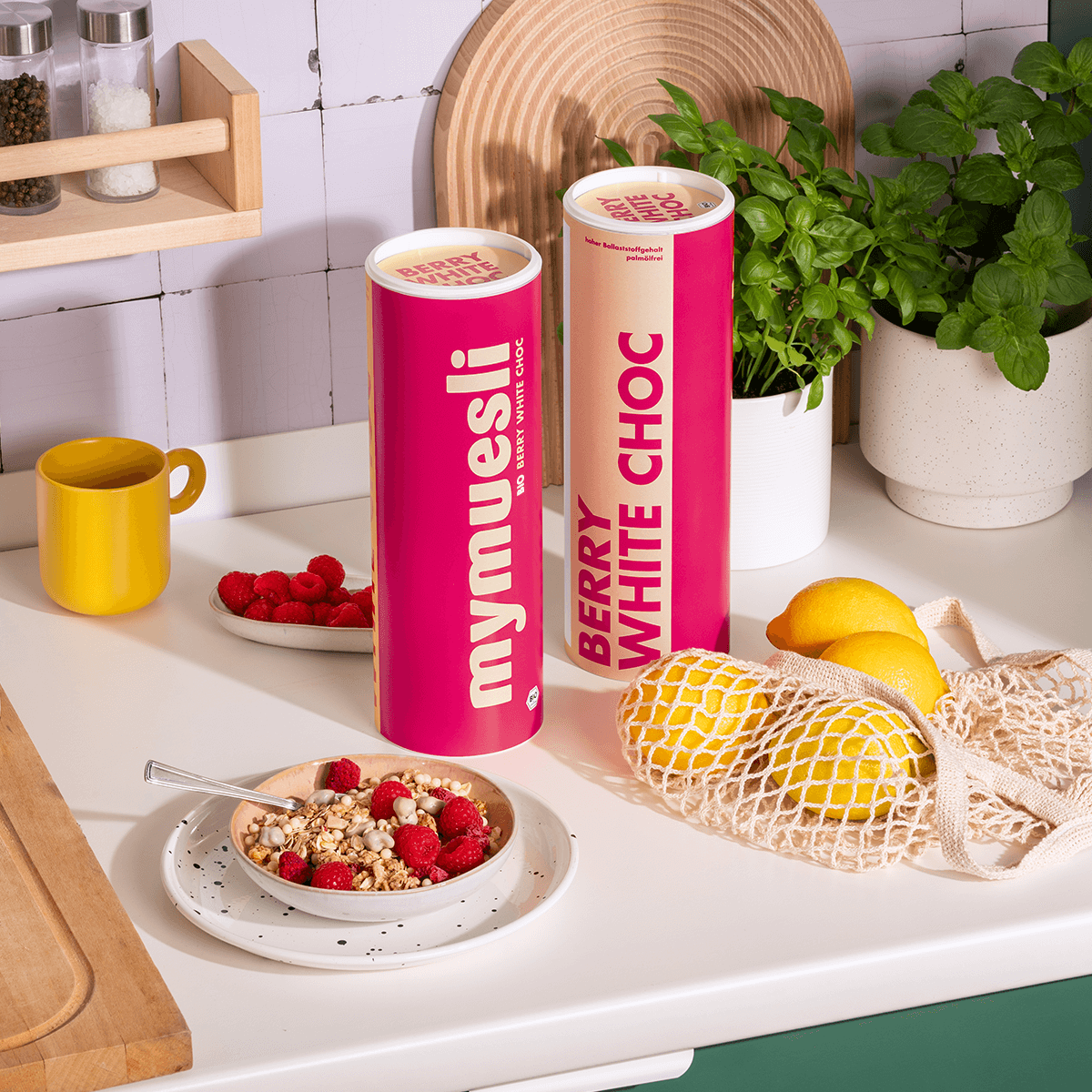 mymuesli Bio Berry White Choc Müsli angerichtet neben Müslidosen, frischen Himbeeren und Zitronen