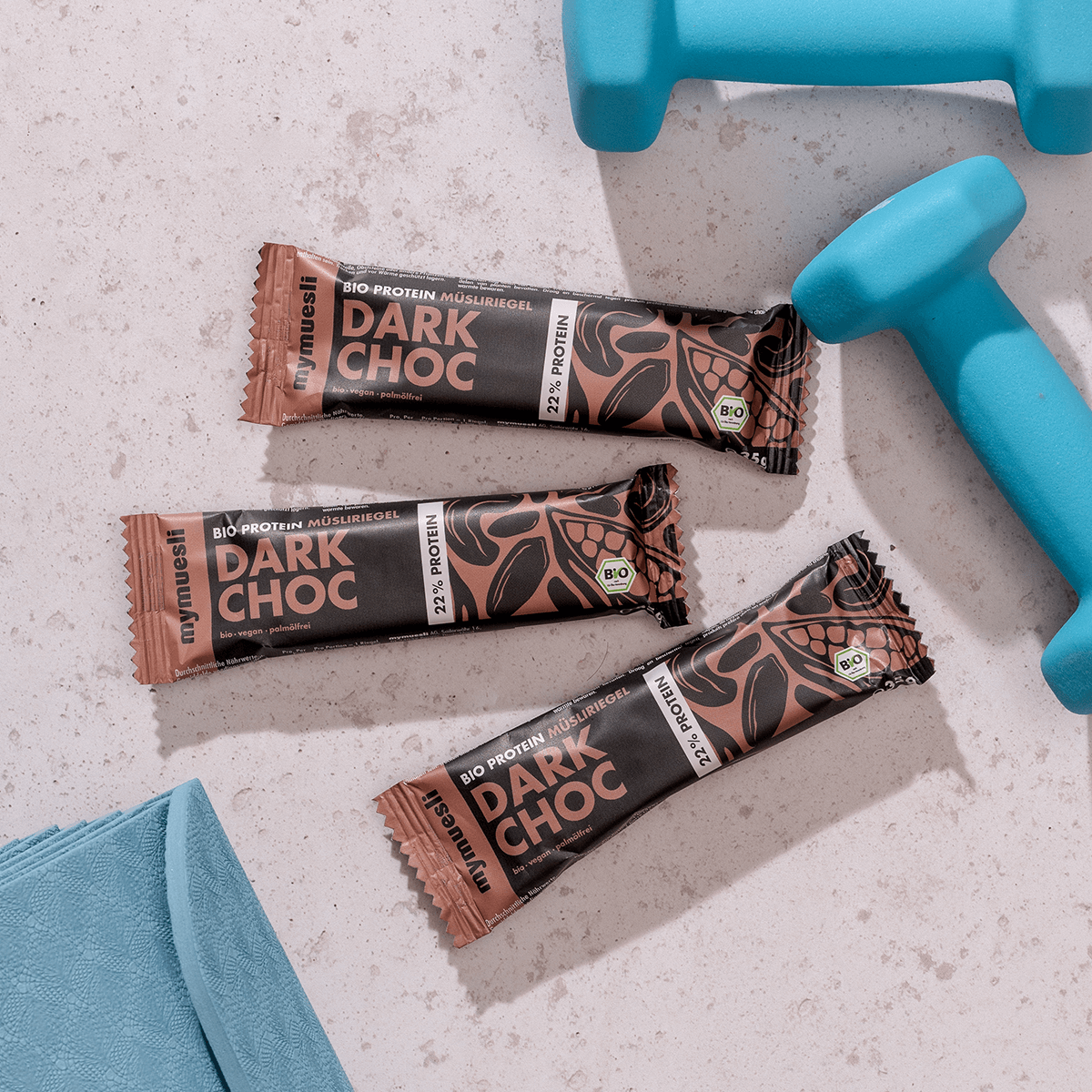 mymuesli Bio Protein Müsliriegel Dark Choc umgeben von Kurzhanteln und Yogamatte.