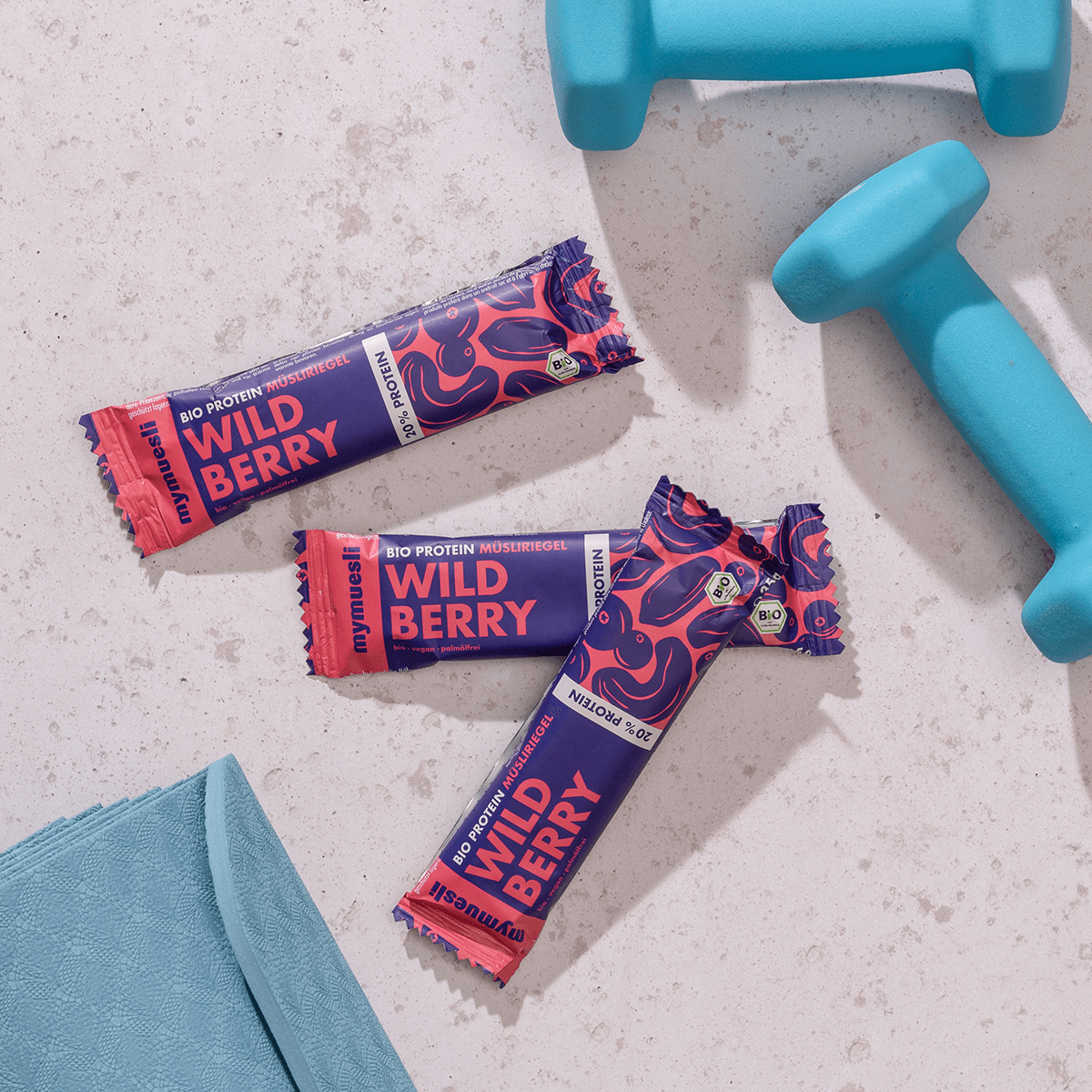 mymuesli Bio Protein Müsliriegel Wild Berry umgeben von Kurzhanteln und Yogamatte.