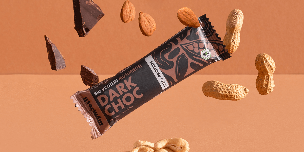 mymuesli Bio Protein Müsliriegel Dark Choc umgeben von Schokolade, Mandeln, Erdnüsse und Cashewkerne.
