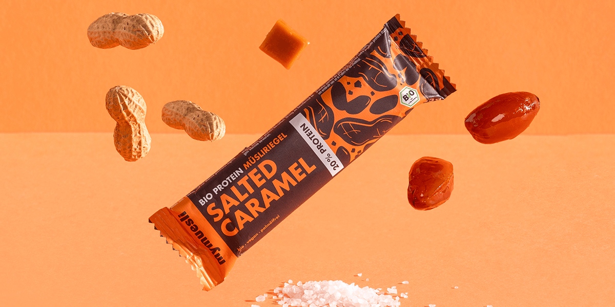 mymuesli Bio Protein Müsliriegel Salted Caramel umgeben von Datteln, Karamell Würfeln und Erdnüssen.
