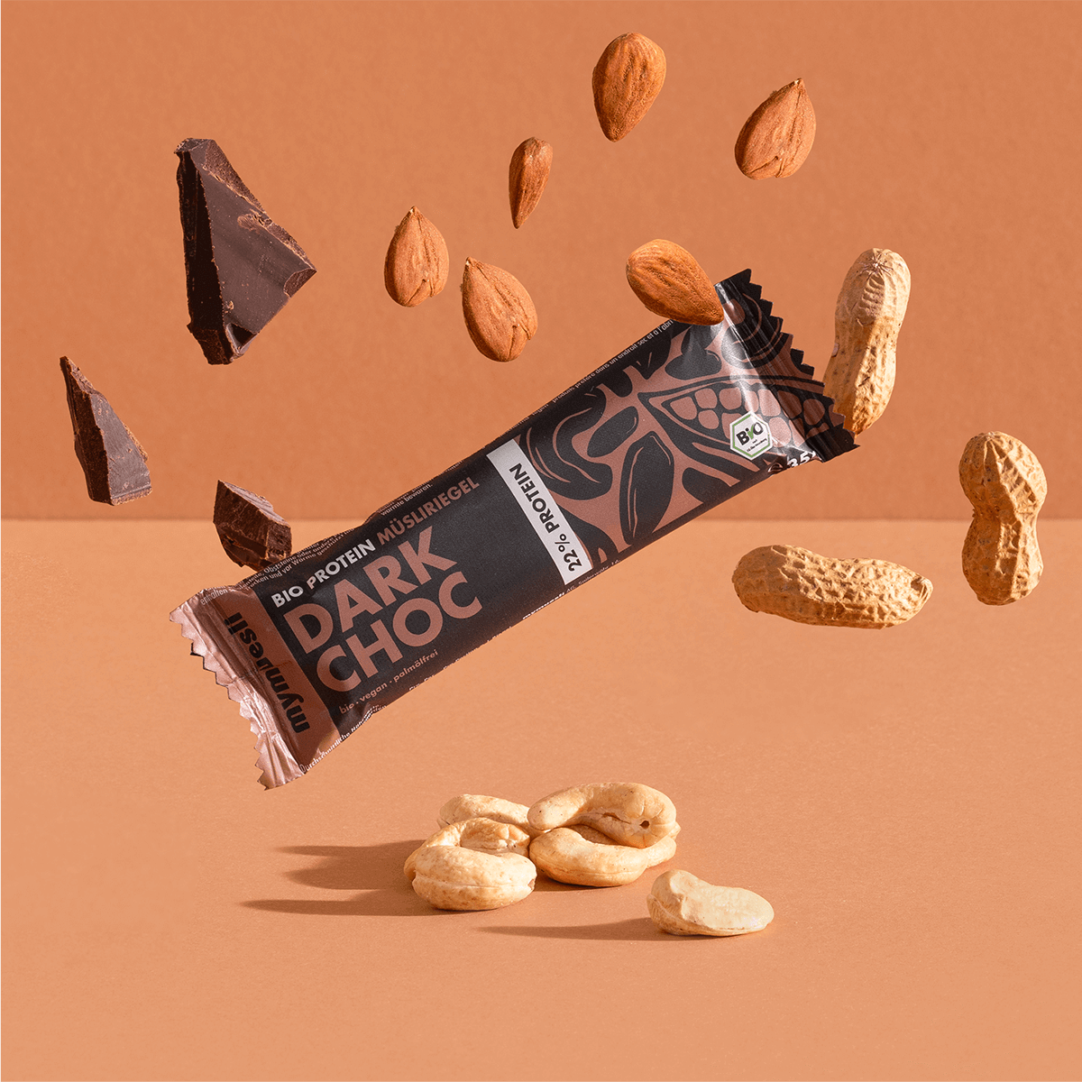 mymuesli Bio Protein Müsliriegel Dark Choc umgeben von Schokolade, Mandeln, Erdnüsse und Cashewkerne.