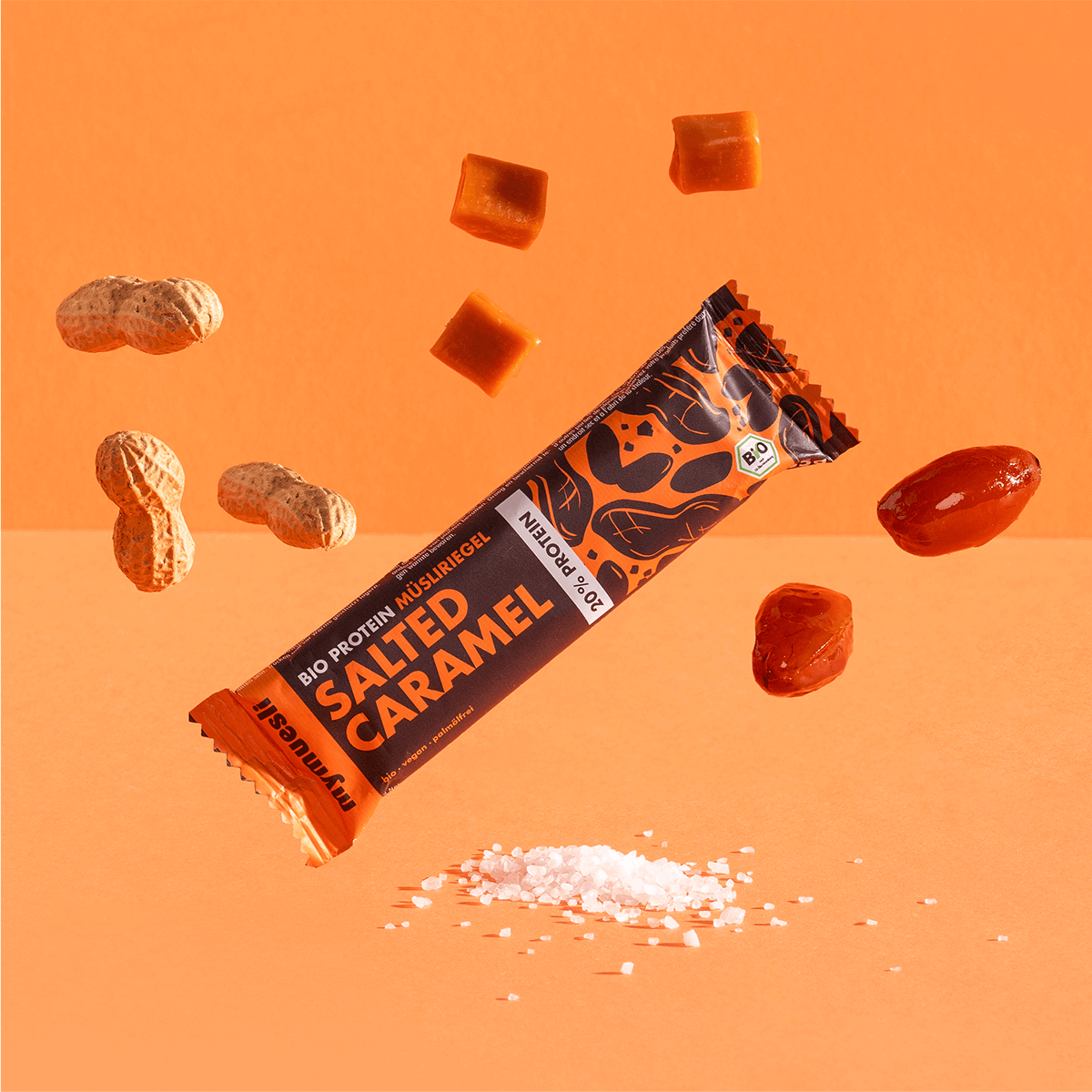 mymuesli Bio Protein Müsliriegel Salted Caramel umgeben von Datteln, Erdnüsse und Karamell Würfel.