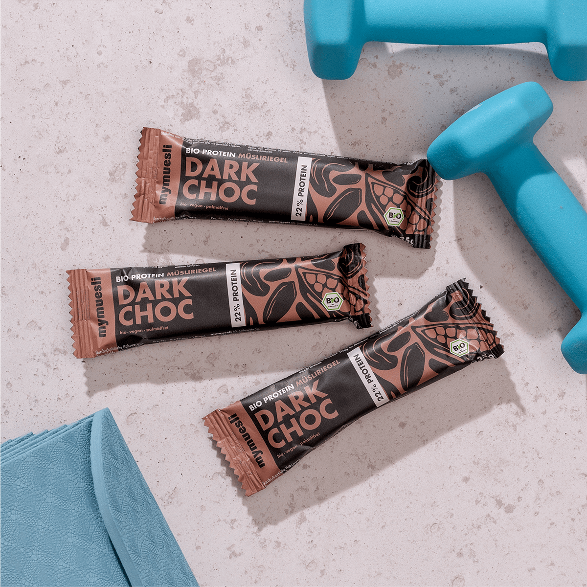 mymuesli Bio Protein Müsliriegel Dark Choc umgeben von frabenfrohen Kurzhanteln und Yogamatte.