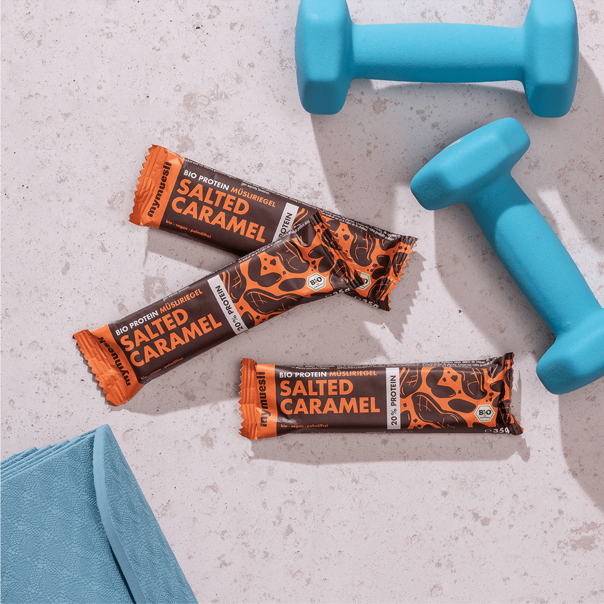 mymuesli Bio Protein Müsliriegel Salted Caramel umgeben von frabenfrohen Kurzhanteln und Yogamatte.