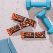 mymuesli Bio Protein Müsliriegel Salted Caramel umgeben von frabenfrohen Kurzhanteln und Yogamatte.