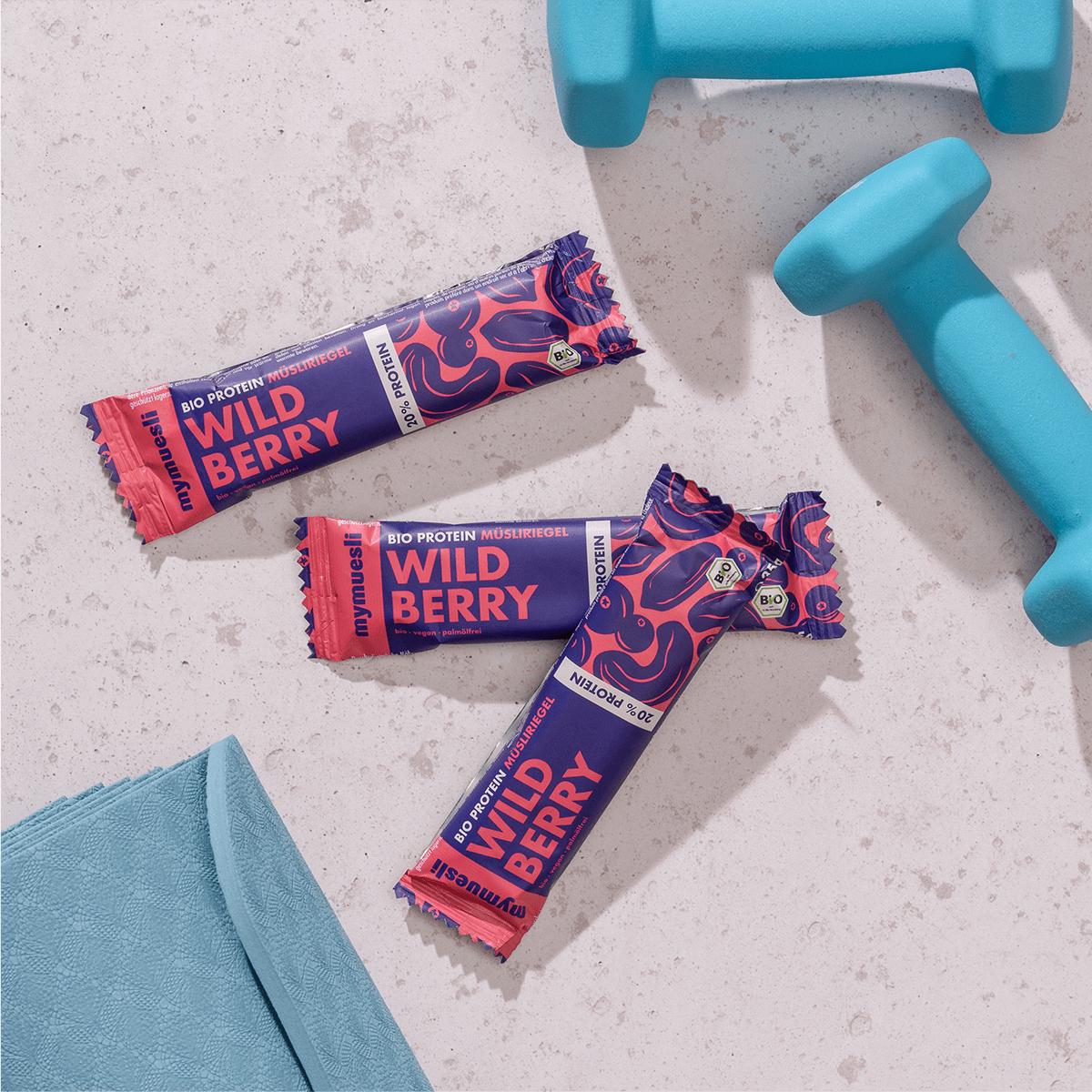 mymuesli Bio Protein Müsliriegel Wild Berry umgeben von frabenfrohen Kurzhanteln und Yogamatte.