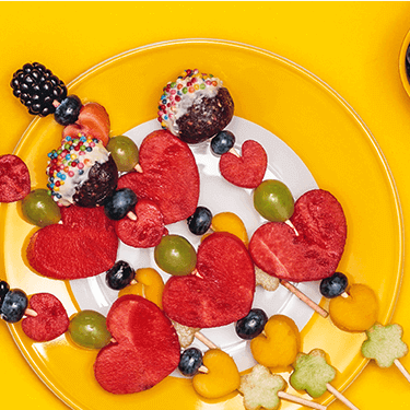 Fruchtspieße für Kinder auf einem Teller mit Weintrauben und Blaubeeren daneben