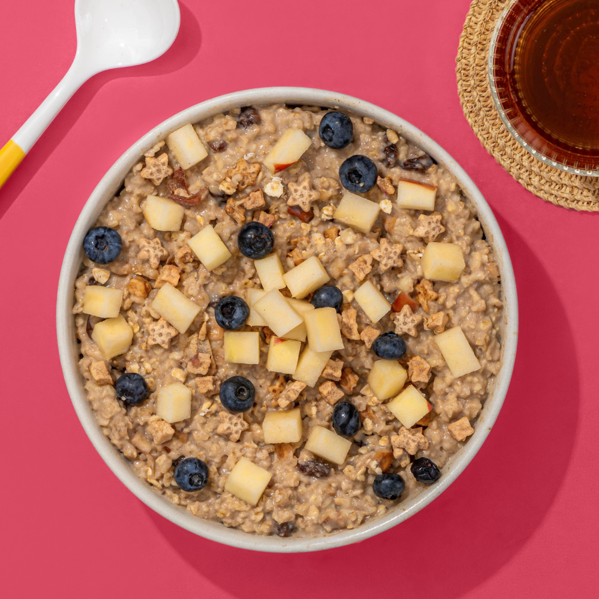 Porridge mit Apfelstücken und Blaubeeren in Müsli Schale, nebendran ein Löffel und ein Glas mit Tee
