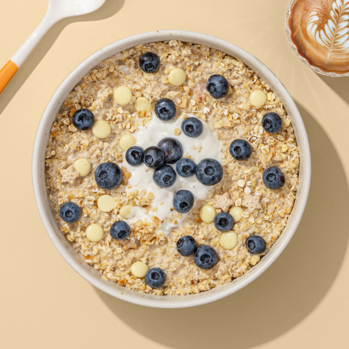 Porridge mit weißen Schokostücken und Blaubeeren, daneben ein Löffel und eine Tasse Kaffee