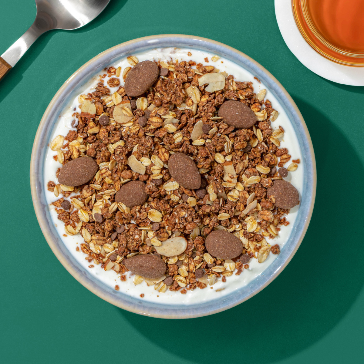 Müsli mit Schokocrunchy, Haferflocken und Schokomandeln in Müslischüssel, nebendran ein Löffel und eine Tasse Tee
