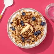 Müsli mit Bratapfelkeksen, Pekannüssen, Blaubeeren und Apfel in Schüssel, nebendran ein Löffel und ein Glas Wasser