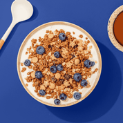 Müsli aus Crunchys, Bratapfelkeksen, Rosinen und Blaubeeren in Müslischüssel, nebendran ein Löffel und eine Tasse Kaffee