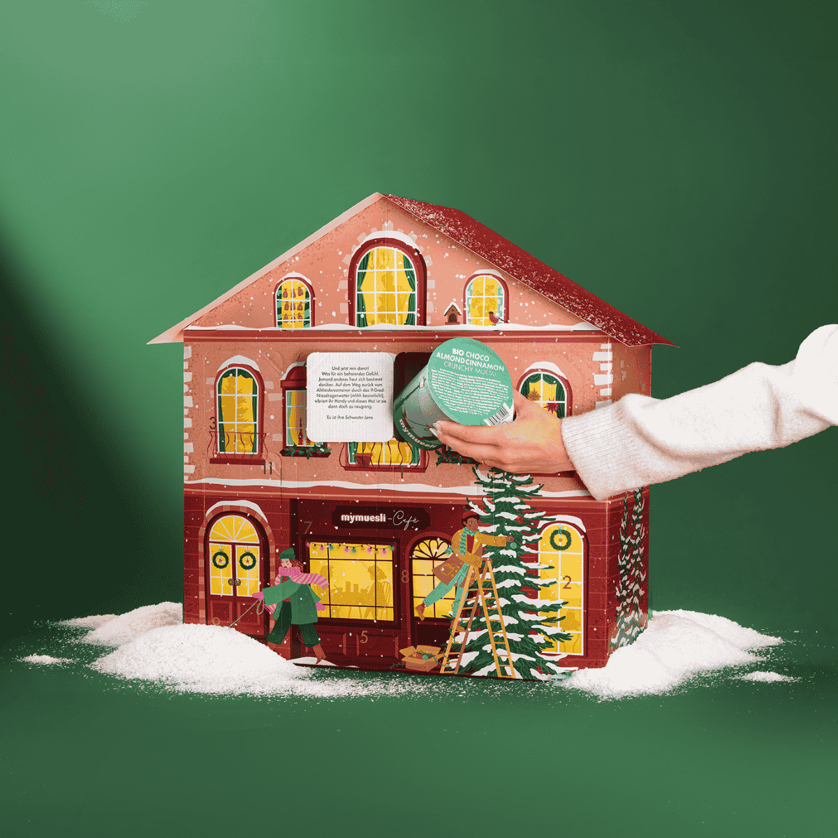 Rotes Adventskalenderhaus mit leichtem Schnee. Vor dem Haus läuft eine Frau mit ihrem Dackel vorbei. Ein Mann steht auf der Leiter und schmückt einen Tannenbaum. Die Fenster des Hauses sind beleuchtet. Eine Frau nimmt einen Müslibecher aus einem geöffneten. 