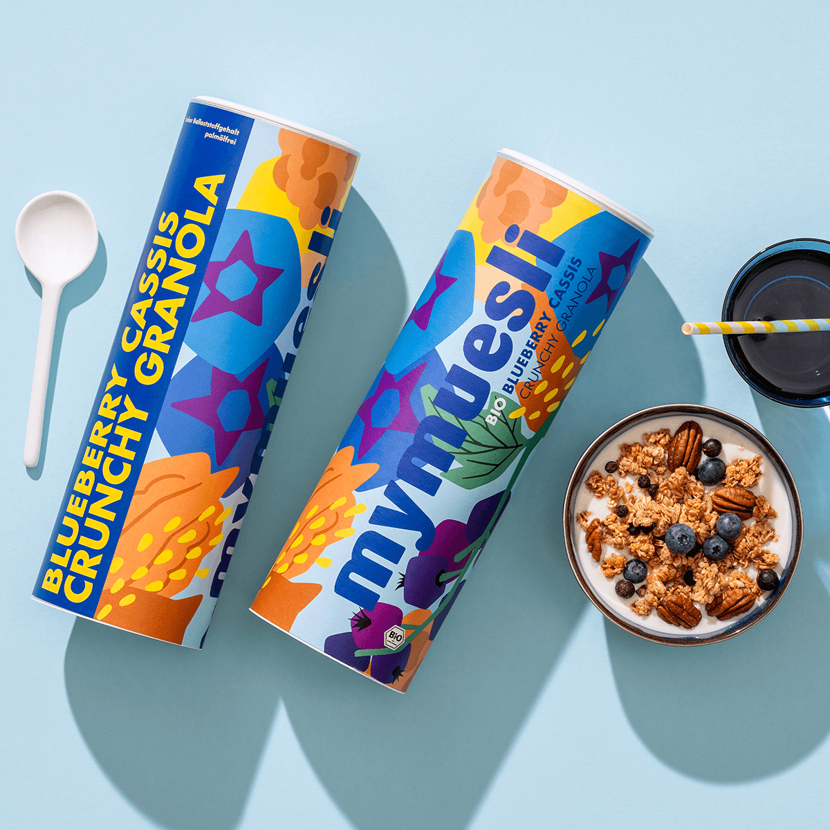 mymuesli Bio Blueberry Cassis Crunchy Granola in Schale neben Smoothie, Dosen und Löffel