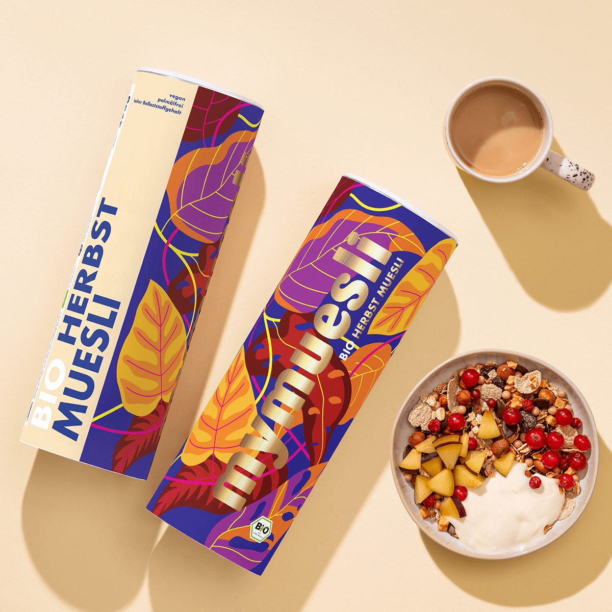 Zwei mymuesli Bio Herbst Müsli Dosen mit herbstlichen Blättern als Design und daneben eine Schüssel Herbst-Müsli mit Johannisbeeren, Pflaumenstücken und Joghurt