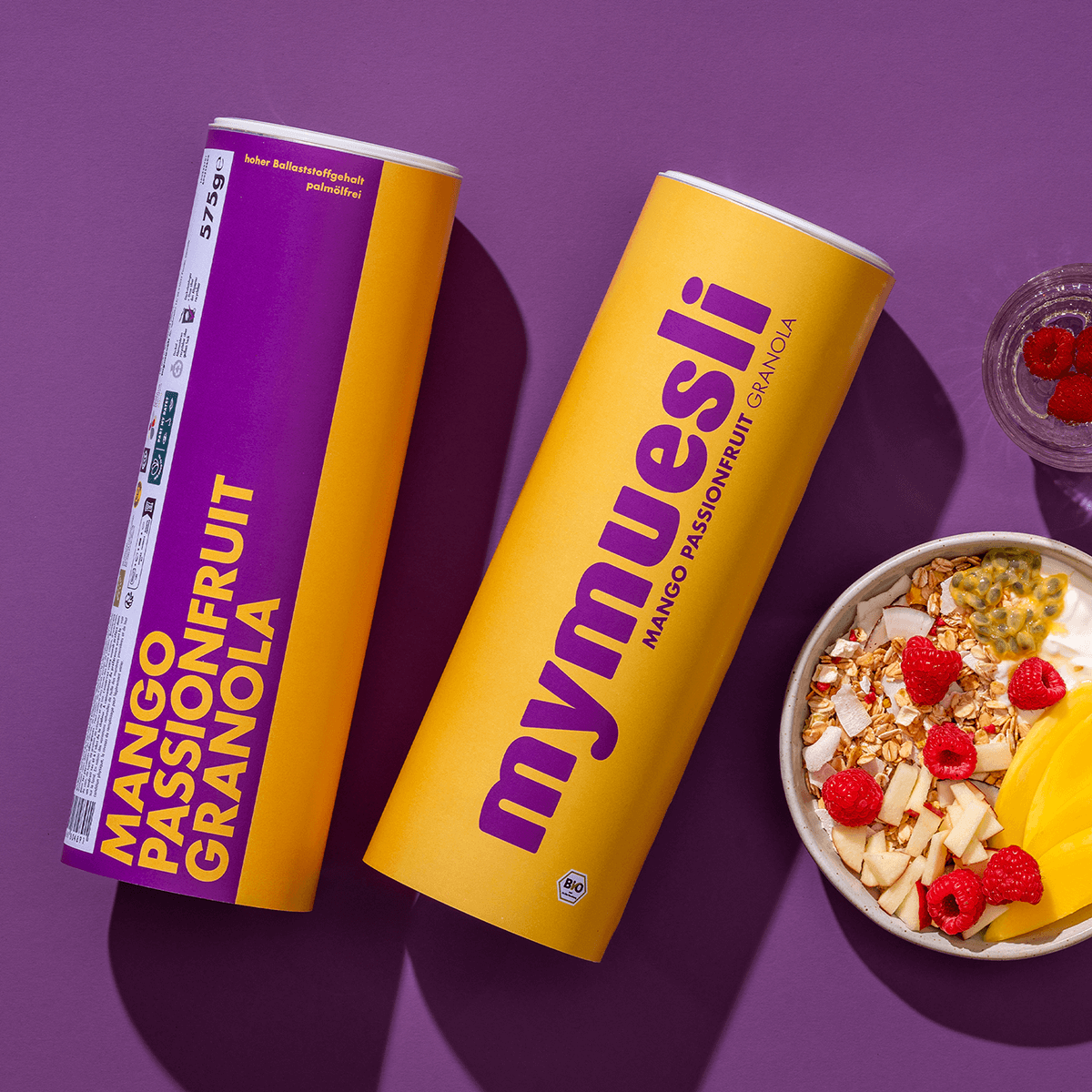 mymuesli Bio Mango Passionfruit Granola Dosen neben Müsli mit Beeren, Mango, Apfel, Passionsfrucht und Kokosnuss