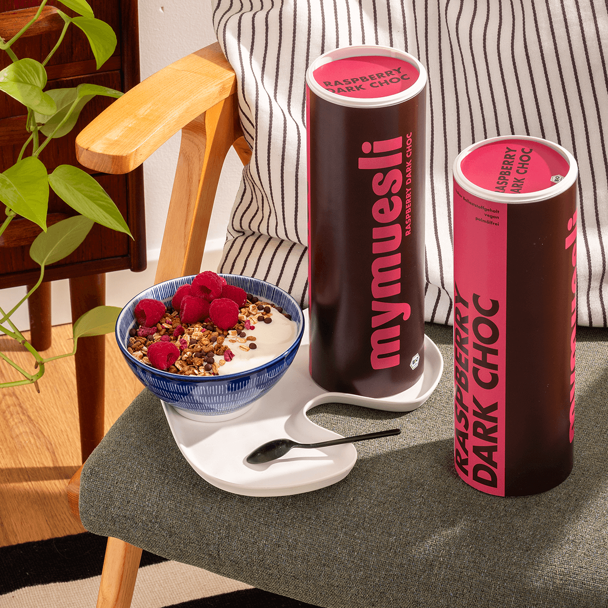 mymuesli Bio Raspberry Dark Choc Müsli in Schale auf Stuhl neben zwei Dosen