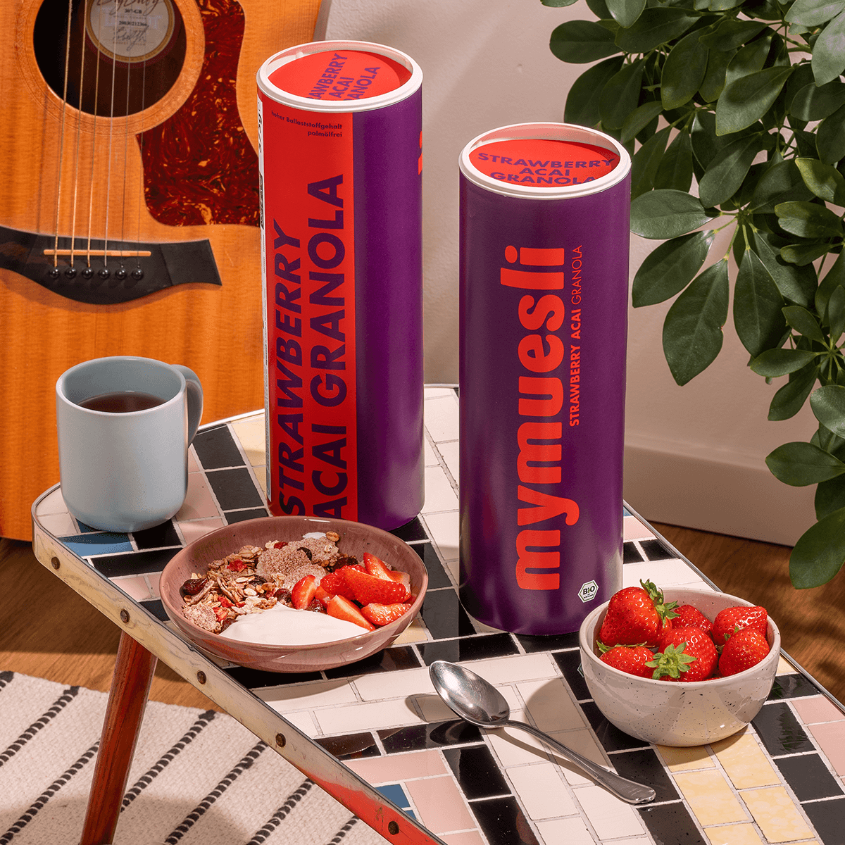 mymuesli Bio Strawberry Acai Granola mit Acai Pulver serviert mit frischen Erdbeeren neben Müsli Dosen