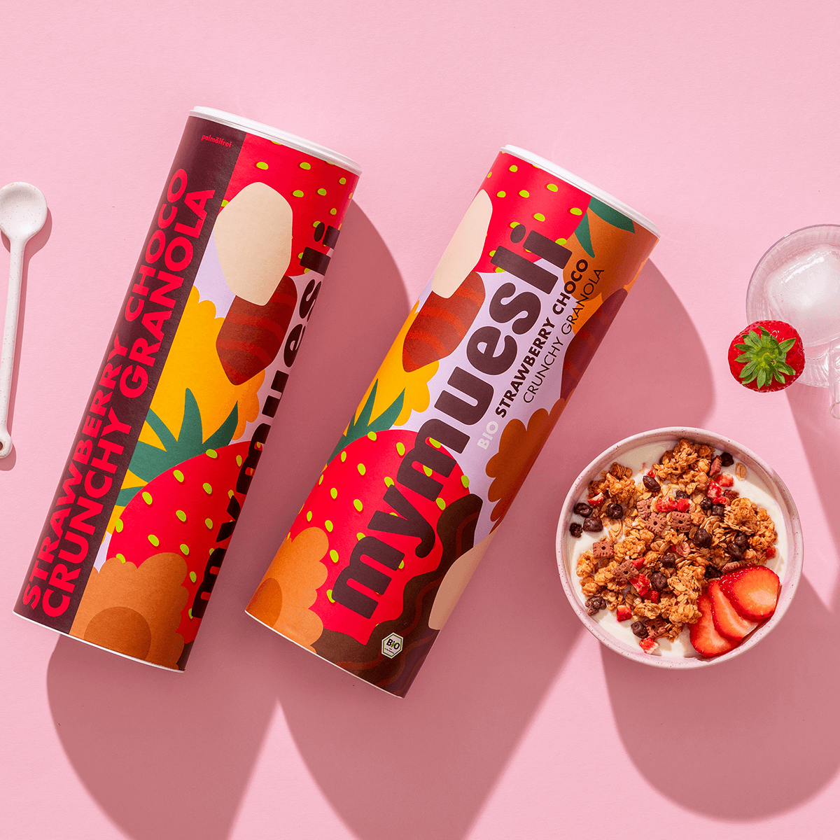 mymuesli Bio Strawberry Choco Crunchy Granola in Schale mit frischen Erdbeeren und Joghurt neben Dosen