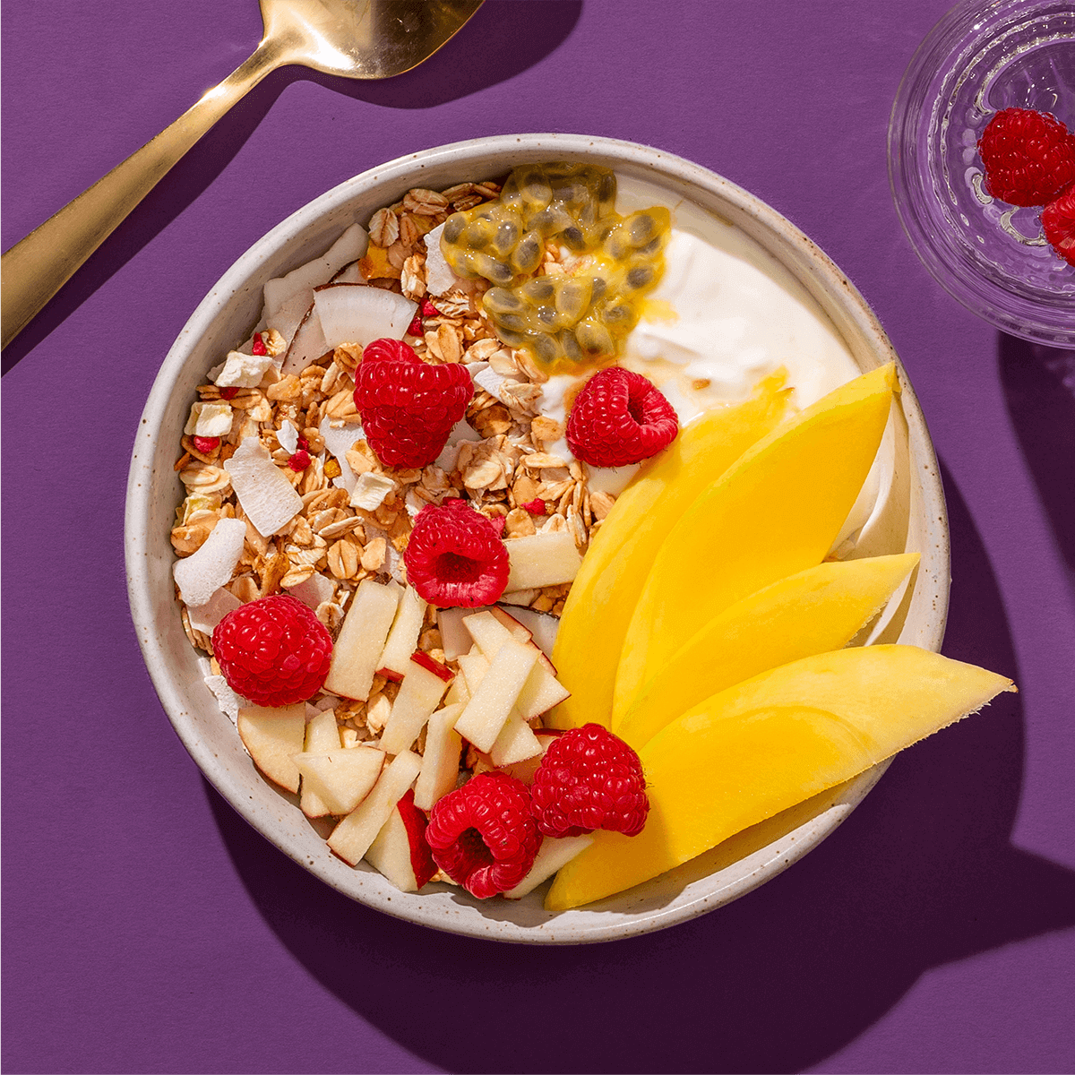 mymuesli Bio Mango Passionfruit Granola in Schüssel mit frischen Himbeeren, Passionsfrucht, Mango, Apfel und Joghurt
