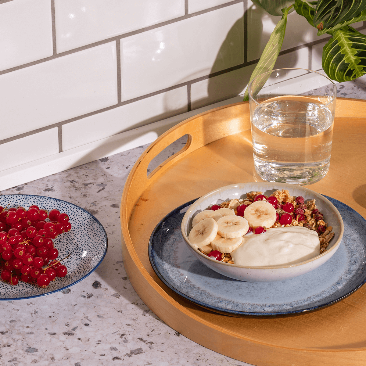 mymuesli Proteinmüsli in Schale mit Bananen und Joghurt auf Tablett mit Wasser neben frischen Johannisbeeren