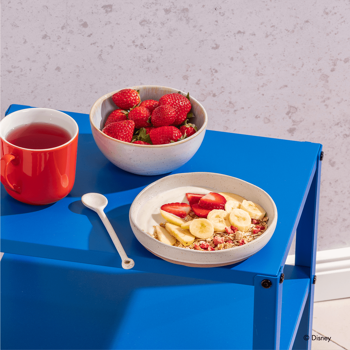 Disney Guten Morgen Löwe Müsli in einer Müslischale mit Bananenscheiben, Erdbeeren, Apfelspalten und Joghurt, daneben eine Schale Erdbeeren und ein Tee