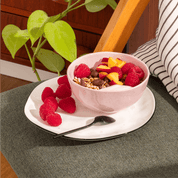 mymuesli Bio Raspberry Dark Choc müsli wird in einer Schale zusammen mit frischen Himbeeren, Pfirsichen und Yoghurt serviert. 