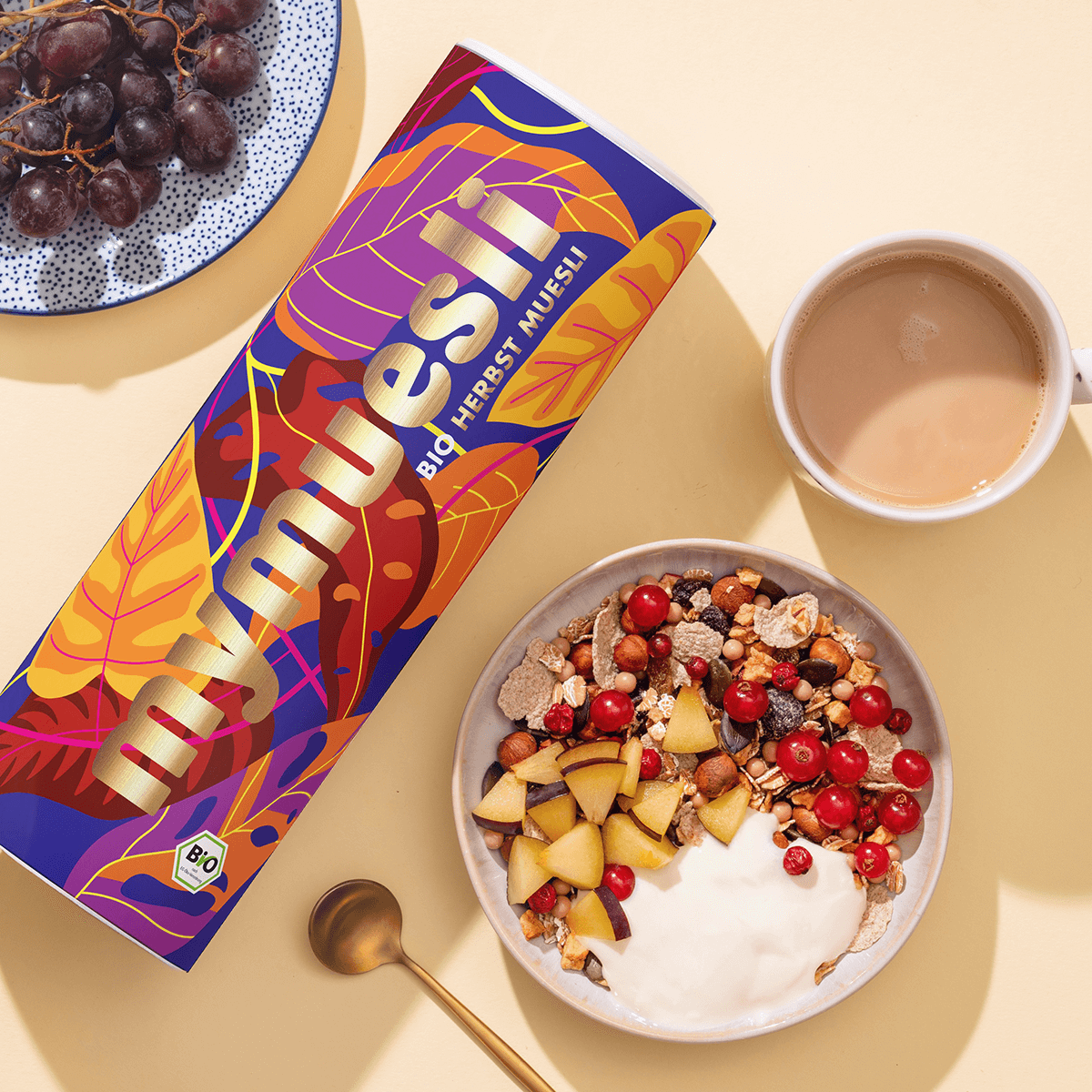 Eine mymuesli Bio Herbst Müsli Dose mit herbstlichen Blättern als Design und daneben eine Schüssel Herbst-Müsli mit Johannisbeeren, Pflaumenstücken und Joghurt
