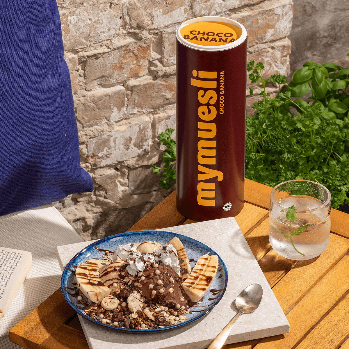 mymuesli Bio Choco Banana Müsli in der Schüssel mit halbierten Bananen, Sahne und Joghurt und dahinter eine Dose Choco Banana Müsli
