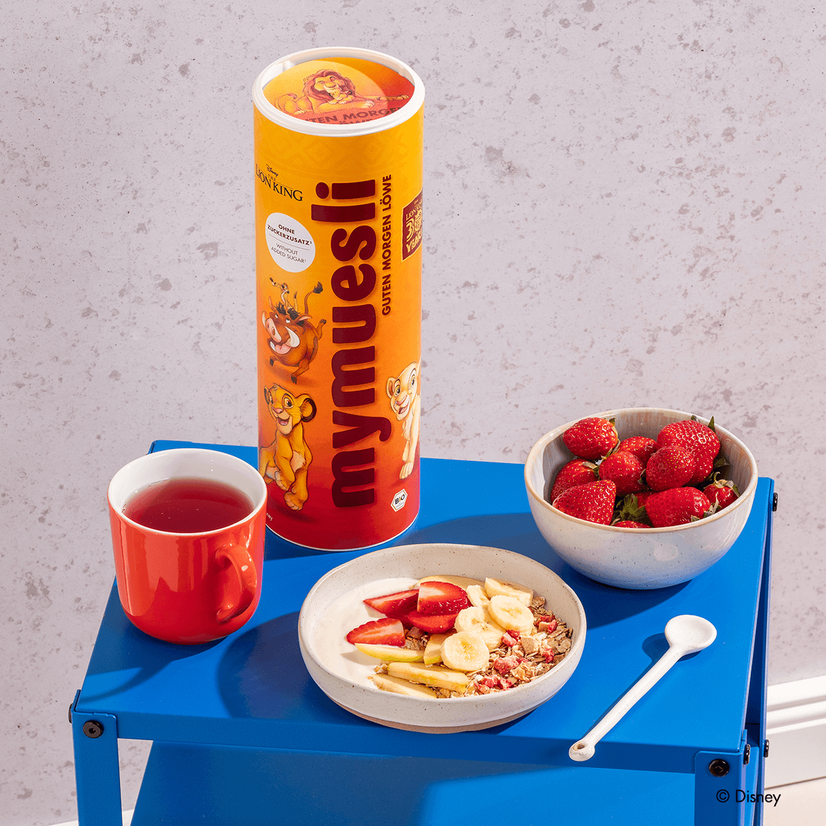Disney Guten Morgen Löwe Müsli Dose, das Müsli in einer Müslischale mit Bananenscheiben, Erdbeeren, Apfelspalten und Joghurt, daneben eine Schale Erdbeeren und ein Tee