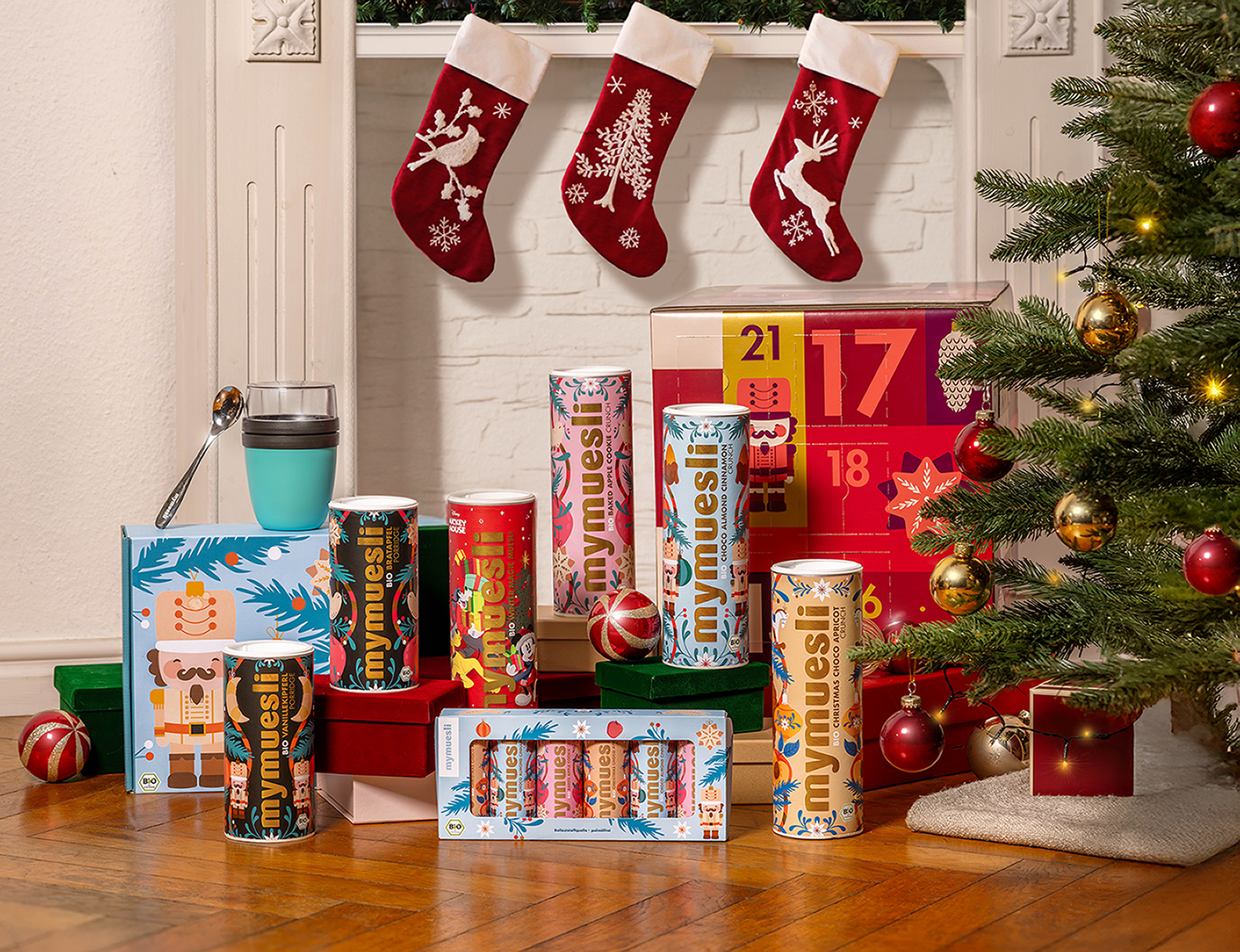 Das Bild zeigt das gesamte mymyuesli Sortiment zu Weihnachten. Dazu gehören: der Müsli Adventskalender, das Christmas Choco Apricot Crunch Müsli, das Baked Apple Cookie Müsli, das Choco Almond Cinnamon Müsli, das Disney Winter Magic Müsli. Dazu gibt es zwei saisonale Porridge in den Richtungen Vanillekipferl und Bratapfel, sowie ein &er Mini Müsli Geschenkset sowie die Müsli Geschenksets mit to Go Becher und silbernen Löffel. Alles steht  zusammen vor einem Kamin, neben dem Weihnachtsbaum.